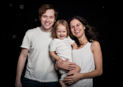 Portrait einer dreiköpfigen Familie. Mutter hält das Baby. Alle haben weißes Oberteil an. Dunkler Hintergrund mit Seifenblasen, die Familie lacht herzlich.