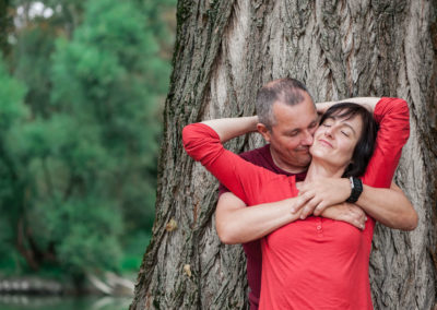 Portrait eines Ehepaars vor einem dicken Baumstamm. Frau steht vor Mann, beide haben die Arme umeinander geschlungen. Im Hintergrund ein See und weitere Bäume.