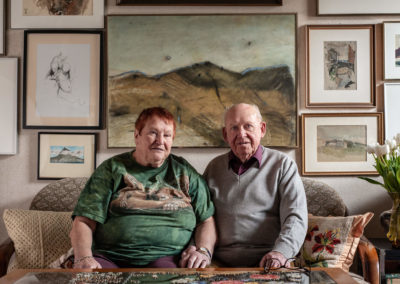 Portrait eines älteren Ehepaares auf ihrem Sofa. Im Hintergrund eine Wand voller Kunstwerke.