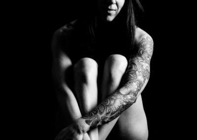 Schwarzweiß-Portrait einer jungen, tätowierten Frau mit langen Haaren. Sie ist Nackt, Beine und Arme sind nah am Körper, mit ihren Händen berührt sie ihre Füße.