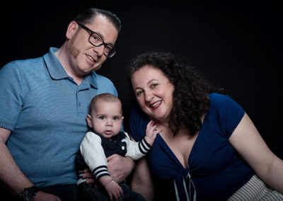 Portait einer dreiköpfigen Familie mit Baby am Schoß des Vaters. Alle in blau gekleidet. Dunkler Hintergrund.
