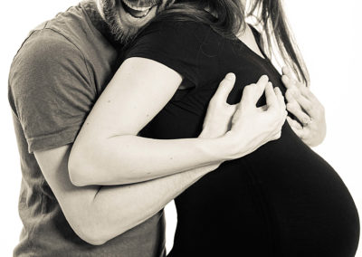 Schwarzweiß-Portrait eines Ehepaars. Frau ist schwanger. Mann umarmt sie von hinten und schaut, verrückt lachend in die Kamera.