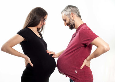 Portrait eines sich gegenüberstehenden Ehepaars. Frau ist schwanger Mann sieht auch so aus. Schauen und greifen sich gegenseitig auf die Bäuche.