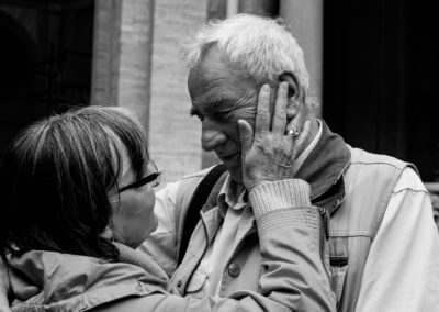Schwarzweiß-Portrait eines älteren Ehepaars vor einem historischen Gebäude. Sie schauen sich tief in die Augen und halten sich inniglich im Arm.