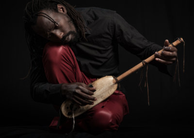 Ein Schwarzer Musiker sitzt verträumt melancholisch mit einem traditionellen Zupfinstrument in dunklem Ambiente.